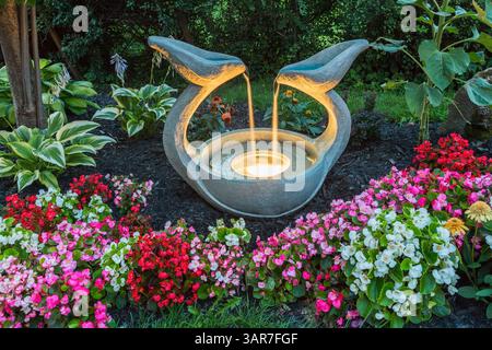 Beleuchtete Wasserfontäne mit Begonia, gelben Chrysanthemen, Echinacea - Coneflower, Hosta in Border im Garten im Hinterhof in der Abenddämmerung im Sommer. Stockfoto