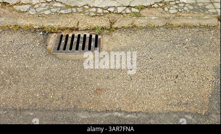 Nahaufnahme eines Kanalrostes, eingebettet in eine zerbröckelnde Asphaltstraße, die den städtischen Verfall und die Infrastrukturprobleme hervorhebt Stockfoto