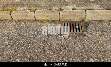 Nahaufnahme eines Kanalrostes, das Wasser auf einer Asphaltstraße neben einer Betonkante ableitet Stockfoto