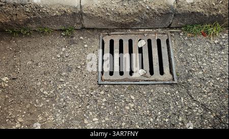 Nahaufnahme eines rostigen Metallgitters, der einen Sturmabfluss auf einer Schotterstraße bedeckt und Anzeichen von Verschleiß zeigt Stockfoto