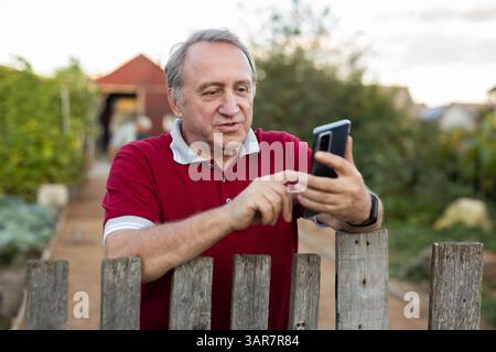 Ein erwachsener Mann, der entspannt neben dem Zaun steht und im Garten telefoniert und lächelt Stockfoto