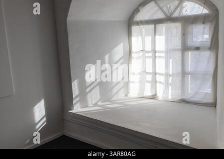 Sonnenlicht strömt durch einen lichtdurchlässigen Vorhang, der ein großes bogenförmiges Fenster in einem makellos weißen Innenraum bedeckt. Das luftige und ruhige Ambiente erinnert an Ruhe Stockfoto