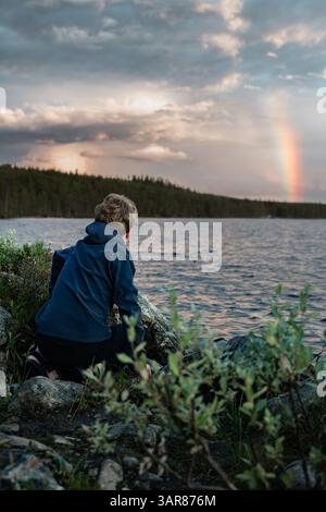 Ein Junge sitzt an Felsen am Wasser, schaut auf den pulsierenden Regenbogen und den Himmel, während der Abend über der ruhigen Landschaft von Dalarna, Schweden, fällt. Stockfoto
