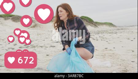 Bild von Social-Media-Herzsymbolen über lächelnder kaukasierin, die Müll vom Strand aufnimmt Stockfoto