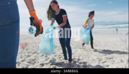 Bild von Herzen über verschiedene weibliche Freiwillige, die Müll am Strand aufsammeln Stockfoto