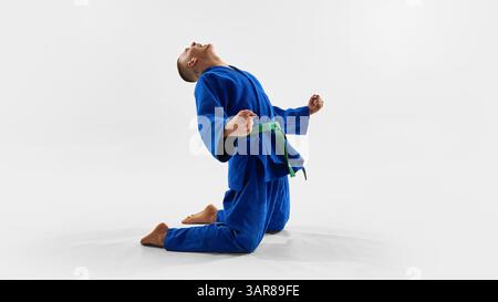 Der Kampfkünstler feiert den Sieg auf Knien, geballte Fäuste, triumphaler Kopfhöhe und drückt emotionale Befreiung, Stolz und Macht im Wettkampfjudo aus Stockfoto