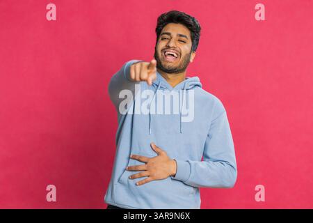 Glücklicher junger indischer Mann, der laut lacht, nachdem er lächerliche Anekdoten gehört hat, Reaktion auf lustigen Witz, unbeschwert amüsiert. Positiver Lebensstil. Arabier isoliert auf rotem Hintergrund Stockfoto
