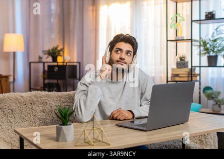 Junger indischer Mann mit schnurlosen Kopfhörern, der am Laptop arbeitet und beim Hörbuch konzentriert wirkt. Der hispanische Kerl sitzt auf dem Sofa am Tisch im Wohnzimmer zu Hause, tief in seine Aufgabe verwickelt. Stockfoto