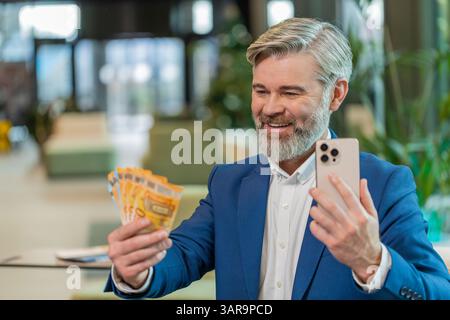 Erfolgreicher glücklicher reifer Geschäftsmann, der ein Smartphone hält und einen Fan von Euro-Rechnungen hält, der in der Lobby des Büros sitzt. Glücklicher kaukasischer bärtiger Kerl, der online Lotteriespiel gewinnt. Budget wird berechnet. Stockfoto