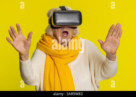 Begeisterte, glückliche Seniorin, die die Headset-Helm-App nutzt, um ein Simulationsspiel zu spielen. 3D 360-Videos in Virtual Reality ansehen. Alte Großmutter in VR-Brille isoliert auf gelbem Wandhintergrund. Zukunftstechnologie Stockfoto