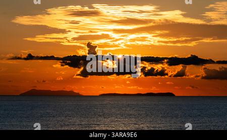 Ein atemberaubender Sonnenuntergang über dem Meer mit leuchtenden Orange- und Gelbtönen, die den Himmel erleuchten. Dunkle Wolken erzeugen einen dramatischen Kontrast, während isla entfernt ist Stockfoto