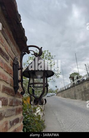 Vintage-Laterne für den Außenbereich an einer Ziegelwand montiert, Kopfsteinpflasterstraße führt bergauf, bewölkter Himmel. Spiegelt rustikalen Charme wider, ideal für Reiseblogs Stockfoto