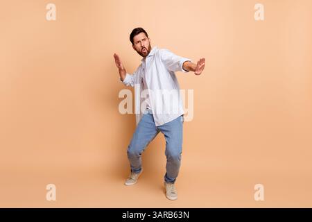 Junger Mann in lässigem Outfit, der eine verspielte Pose auf beigefarbenem Hintergrund macht, ausdrucksstarke Handgesten macht und trendigen Lifestyle zum Ausdruck bringt Stockfoto