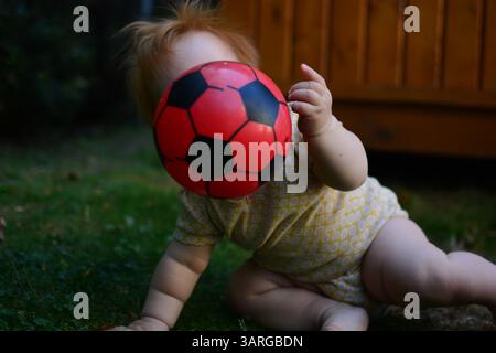 Rotes Haar Baby Mädchen in gelbem Body spielt in einem Hinterhof mit rotem und schwarzem Fußball. Kleinkind Mädchen sitzt draußen auf Gras und wirft b Stockfoto