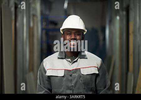 Porträt eines jungen afroamerikanischen Vorarbeiters in grauer Uniform und mit Helm auf dem Kopf, der in die Kamera lächelt Stockfoto