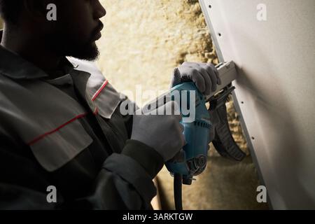 Mittlere Nahaufnahme eines nicht erkennbaren afroamerikanischen männlichen Baumeisters in grauer Uniform, der die Nagelpistole hält, während er den Nagel in die Oberfläche treibt Stockfoto
