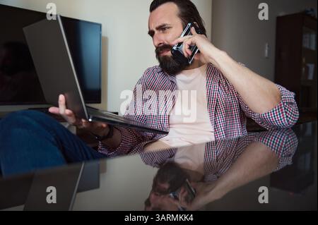 Zielgerichteter junger Mann, der Aufgaben auf einem Laptop verwaltet, während er im Home Office über ein Smartphone spricht. Stockfoto