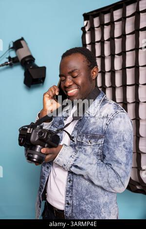 Afroamerikaner, der auf den Kamerabildschirm schaut, Bilder durchliest und mit dem Kunden im Studio telefoniert. Ein schwarzer Fotograf hält das Handy am Ohr und stöbert gerne in seiner Fotogalerie. Stockfoto