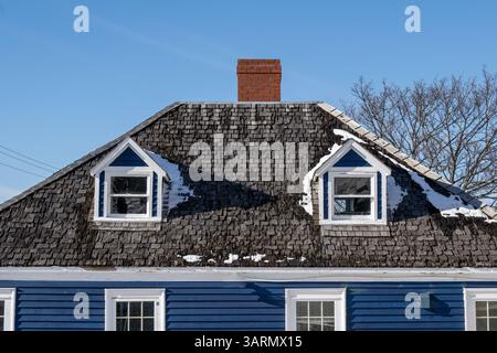 Klassisches Dach im Mansardenstil mit zwei Dachfenstern, Zedernweindach, rotem Ziegelkamin, marineblauer horizontaler Schranke und doppelt hängenden Fenstern. Stockfoto