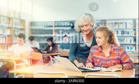 Eine freundliche Lehrerin hilft Schülerinnen, Informationen auf einem Laptop in der Schulbibliothek zu finden Stockfoto