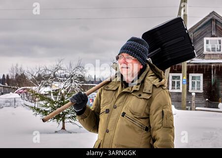 Individual in olivgrüner Winterbekleidung hält schwarze Schneeschaufel während der kalten Jahreszeit mit rustikaler Landschaft und schneebedeckter Landschaft Stockfoto