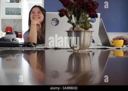 Eine junge Frau lächelt, während sie eine Pause von der Arbeit oder dem Lernen auf ihrem Laptop an einem reflektierenden Tisch einlegt. Sie ist zu Hause. Stockfoto