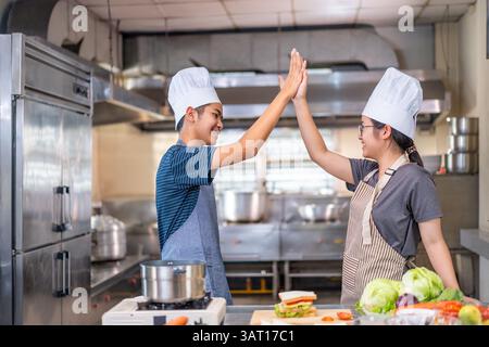 Zwei junge Köche feiern mit einem High-Five in einer kommerziellen Küche, tragen Kochmütze und Schürzen und zeigen Teamarbeit und Freude beim Kochen. Stockfoto