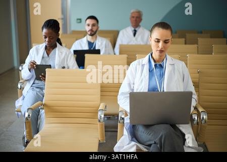 Gruppe medizinischer Fachkräfte, die sich mit ihren Laptops im Wartezimmer des Krankenhauses beschäftigen. Die Szene umfasst ein vielfältiges Team mit kaukasischen und afroamerikanischen Doktoren Stockfoto