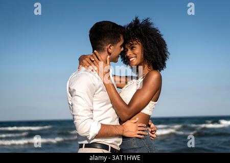 Glückliches junges Paar, das sich während der goldenen Stunde an einem Strand umgibt, lächelt und einen romantischen Sommermoment am Meer genießt. Stockfoto