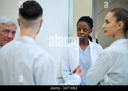 Multiethnische Ärztegruppe diskutiert Patientenbehandlungsplan im Krankenhaus. Das Team umfasst leitende Ärzte und junge Fachkräfte in medizinischen Uniformen, Stockfoto