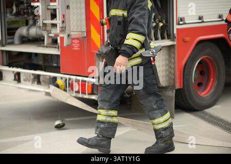Ein Feuerwehrmann in Vollweichenausrüstung geht neben einem aktiven Feuerwehrfahrzeug mit einem Schlauch auf dem Boden und zeigt mit sichtbaren Werkzeugen die Notfallbereitschaft, Sicherheit Stockfoto