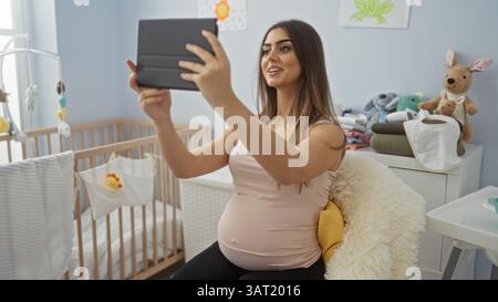 Schwangere Frau macht Selfie in einem gemütlichen Babyzimmer mit Wiege, umgeben von weichen Spielzeugen und wichtigen Babysachen, die einen fröhlichen Moment in der Wohnung festhalten. Stockfoto