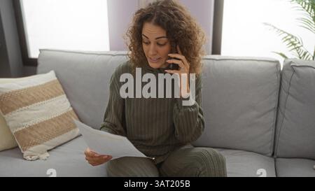 Frau im Wohnzimmer telefoniert, lächelt beim Lesen von Dokumenten, entspannt auf dem Sofa, entspannt in gemütlicher Apartmentatmosphäre, mit modernem Lebensstil Stockfoto