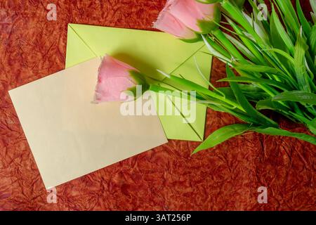 Eine rosafarbene Rose in einem grünen Umschlag, platziert auf einem strukturierten Stoff mit einer leeren Karte, ist perfekt für Gruß- oder Kommunikationsthemen. Stockfoto