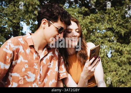 Ein junges Paar, das im sonnigen Park glücklich auf das Telefon schaut Stockfoto
