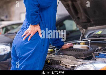 Rückansicht eines nicht erkennbaren Mannes mit Mechaniker, der seinen unteren Rücken aufgrund von Schmerzen in der Werkstatt unangenehm hält Stockfoto