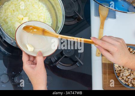 Eine Hand, die einen Holzlöffel über gebratenen Zwiebeln in einem Topf hält. Gehackte Pilze, Kartoffeln und ganze Pilze auf der Seite. Hausmannskost läuft, bis Stockfoto