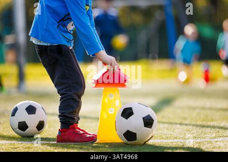 Ein Kind beim Fußballtraining legt rote Kegel auf eine gelbe Markierung mit zwei Fußballbälle in der Nähe auf einem sonnigen Feld. Fußball-Trainingsfeld. Andere Kinder spielen mich Stockfoto