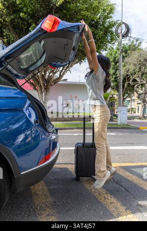 Ein Teenager steht neben einem blauen Auto und zieht einen Koffer raus. Die Szene ist lässig und entspannt, die Frau bereitet sich wahrscheinlich auf eine Reise vor Stockfoto