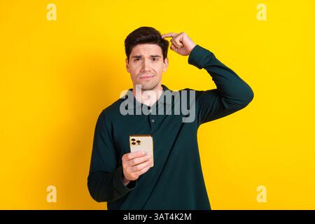 Junger Mann in grünem Hemd mit einem Telefon, das verwirrt aussieht, während er den Kopf kratzt, vor leuchtendem gelbem Hintergrund Stockfoto