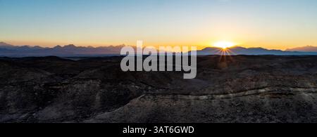 Ein atemberaubender Sonnenaufgang über einer zerklüfteten Wüstenlandschaft mit Bergen im Hintergrund und einem klaren Himmel. Die Sonnenstrahlen platzen durch den Horizont, krank Stockfoto