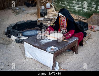 Eine Frau in traditioneller Kleidung rollt Teig auf einem Holztisch in einer sandigen Umgebung im Freien. Der Hintergrund zeigt eine Feuerstelle und rustikale Elemente, c Stockfoto