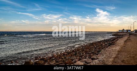 Ein ruhiger Blick auf die Küste mit einer felsigen Küste, sanften Wellen und einem klaren Himmel mit schimmernden Wolken. Die Sonne untergeht und strahlt ein warmes Licht über die W Stockfoto
