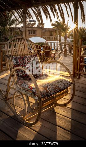 Ein rustikaler Schaukelstuhl mit einem bunten Kissen sitzt auf einer Holzterrasse, die in warmes Sonnenlicht getaucht ist. Palmen und eine gemütliche Atmosphäre schaffen ein ruhiges Outdoo Stockfoto