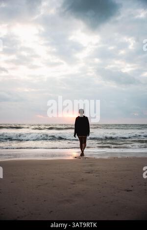 Frau mit Blick auf das Meer bei Sonnenuntergang, barfuß am Strand mit dramatischen Wellen und einem leuchtenden Horizont. Stockfoto