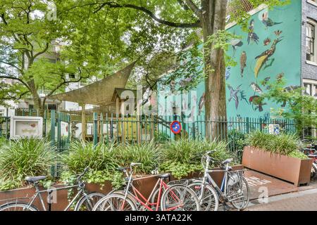 Eine lebhafte urbane Parkszene mit üppigem Grün, einem einzigartigen Wandgemälde am Gebäude und geparkten Fahrrädern, die eine lebhafte Atmosphäre im Freien schaffen. Stockfoto