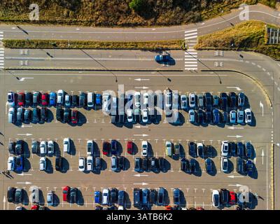Aus der Vogelperspektive auf einen großen, organisierten Parkplatz voller farbenfroher Autos. Helles Sonnenlicht wirft lange Schatten und hebt die ordentlichen Reihen und Straßenmarkierungen hervor Stockfoto