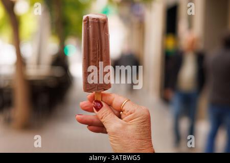 Billiges Schokoladeneis auf einem Stock, der von der Hand eines nicht erkennbaren älteren Menschen gehalten wird Stockfoto