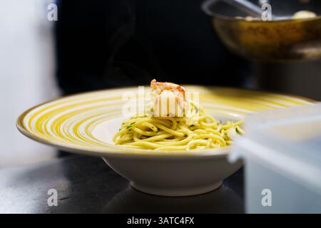 Eine Dampfplatte mit Garnelen-Spaghetti-aglio e olio wird kunstvoll auf einem gelb gestreiften Gericht präsentiert. Die Nudeln sind mit Olivenöl überzogen und garniert Stockfoto