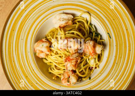 Ein wunderschön überzogener Garnelen-Spaghetti-aglio e olio wird auf einer gelb gestreiften Keramikschale auf einem Holztisch präsentiert. Die Teigwaren sind mit Olivenöl überzogen Stockfoto